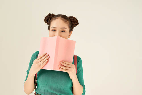 Charming female  and hiding half of face behind book with pink cover while standing on light background