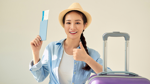 Indoor shot of young attractive traveler tourist woman wearing summer casual clothes, posing with suitcase, tickets and passport, showing thumb up isolated over gray wall. Passenger traveling abroad.