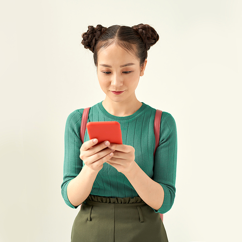 A portrait of an attractive young asian woman carrying backpack standing isolated over white background, using mobile phone