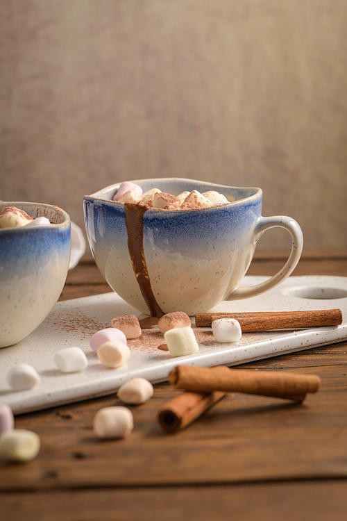 Cups of hot chocolate drink with marshmallows and cinnamon on brown wooden background.
