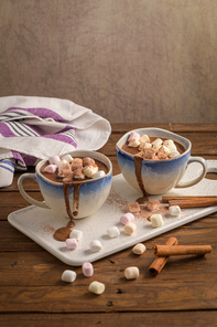 Cups of hot chocolate drink with marshmallows and cinnamon on brown wooden background.