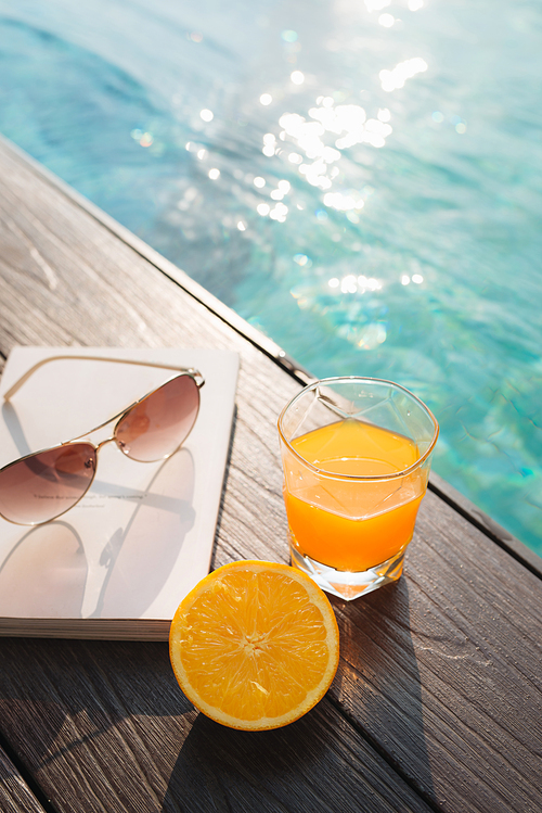 Orange fresh juice, book and sunglasses near pool
