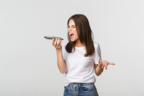 Angry, pissed-off brunette girl arguing, record voice message on mobile phone, white background.