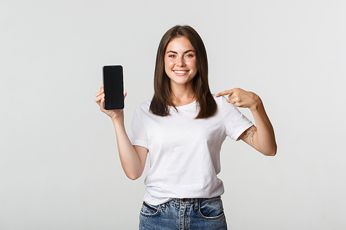Attractive smiling woman pointing finger at smartphone screen, white background.