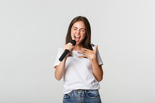 Carefree beautiful girl singing karaoke, holding microphone, standing white background.