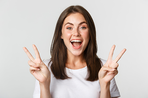Portrait of beautiful and happy brunette girl smiling and showing peace gestures, standing white background excited.