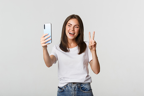 Attractive brunette girl looking happy and taking selfie on smartphone, showing peace gesture.