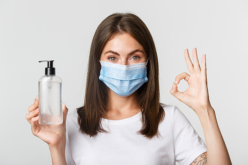 Covid-19, health and social distancing concept. Close-up of smiling brunette girl in medical mask, showing hand sanitizer and okay gesture.