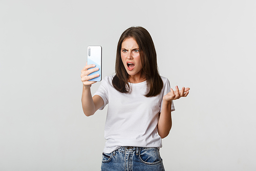 Frustrated young woman having argument on video call, holding smartphone, standing white background.