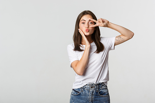 Beautiful young woman showing peace gesture and pouting silly, white background.