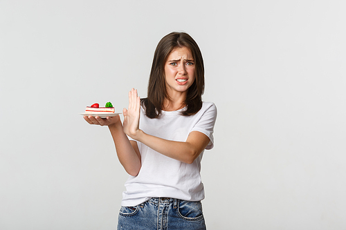 Disgusted young woman rejecting eating awful cake, making refusal gesture and grimacing displeased.