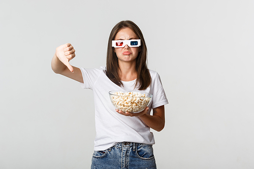 Disappointed attractive girl in 3d glasses, holding popcorn and showing thumbs-down, dislike movie.