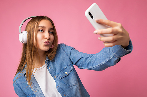 attractive happy smiling young blonde woman wearing blue jean stylish shirt and casual white t-shirt isolated over pink background wearing white bluetooth wireless headphones and listening to good music and using mobile phone having video call communication looking at gadjet