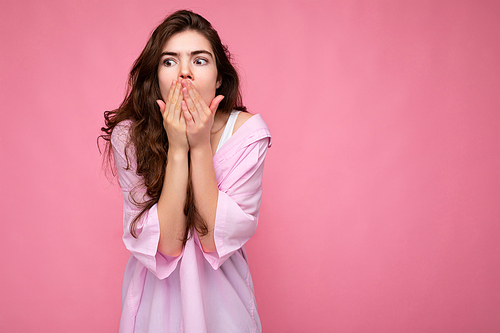 Photo of young shocked beautiful curly brunette woman with sincere emotions wearing casual pink shirt isolated over pink background with copy space and covering mouth with hands.