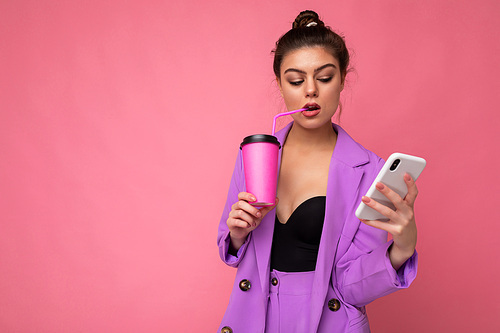 Shot of concentrated caucasian brunette woman holds take away coffee dressed in casual clothes isolated over pink wall uses smartphone and drinking.