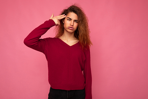 Portrait of young emotional beautiful brunette curly woman with sincere emotions and grimace wearing trendy pink pullover isolated on pink background with copy space and showing gun gesture with hand.