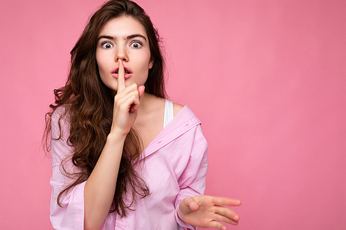 Photo of young shocked beautiful wavy-haired brunette woman with sincere emotions wearing casual pink shirt isolated over pink background with copy space and showing silence gesture with finger near lips. Shhh sign.
