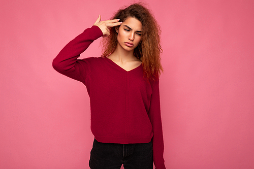 Portrait of young emotional tired beautiful brunette curly woman with sincere emotions wearing trendy pink pullover isolated on pink background with copy space and showing gun gesture with hand.