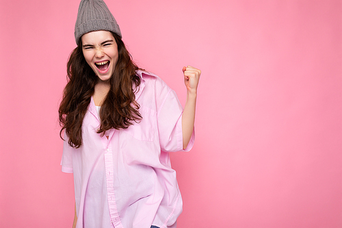 Attractive positive happy young curly brunette woman wearing pink shirt and gray hat isolated on pink background with copy space and celebrating victory.