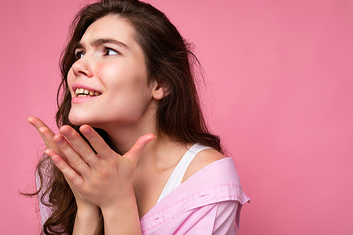 Closeup of young shocked amazed beautiful curly brunette woman with sincere emotions wearing casual pink shirt isolated over pink background with copy space and looking to the side.