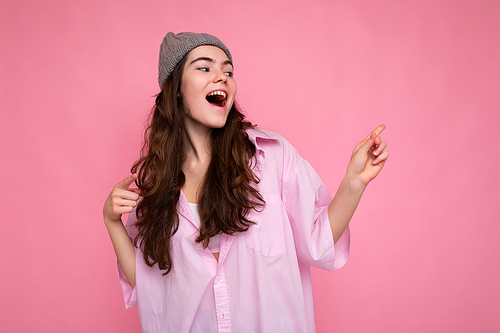 Beautiful happy positive joyful young curly brunette woman wearing pink shirt and grey hat isolated on pink background with copy space.