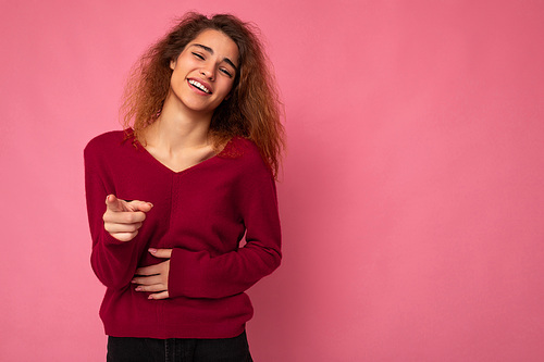 Young european happy positive charming cute brunette curly woman with sincere emotions wearing trendy pink sweater isolated over pink background with free space and laughing.