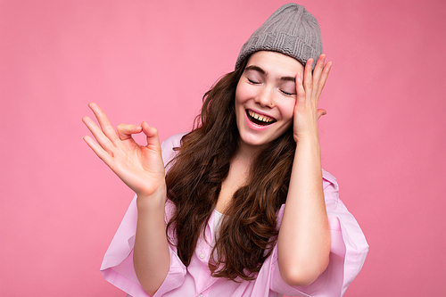 Portrait of young positive happy beautiful curly brunette woman with sincere emotions wearing trendy pink shirt and grey hat isolated on pink background with empty space and showing ok gesture. It's good.