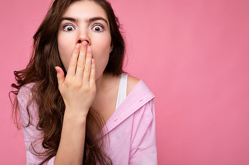 Photo of young shocked beautiful wavy-haired brunette woman with sincere emotions wearing casual pink shirt isolated over pink background with copy space and covering mouth with hand.