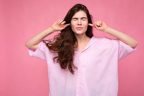 Portrait of young emotional winsome nice curly brunette woman with sincere emotions wearing casual pink shirt isolated over pink background with copy space and covering ears with fingers and closed eyes.