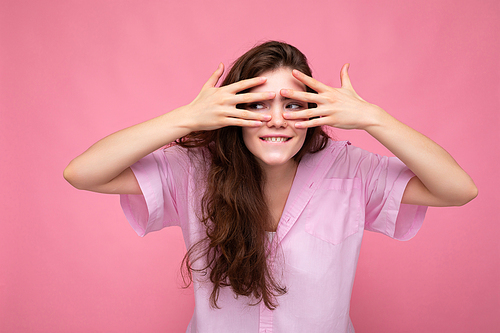 Portrait of young emotional positive funny beautiful curly brunette woman with sincere emotions wearing casual pink shirt isolated over pink background with copy space and covering eyes with hands and having fun.