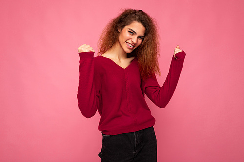 Young emotional happy positive beautiful brunette curly woman with sincere emotions wearing casual pink pullover isolated over pink background with copy space and celebrating winning and raising fists.