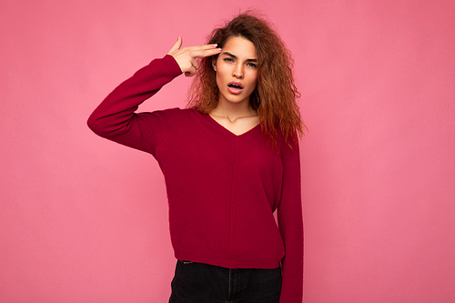 Portrait of young emotional beautiful brunette curly woman with sincere emotions wearing trendy pink pullover isolated on pink background with copy space and showing gun gesture with hand.