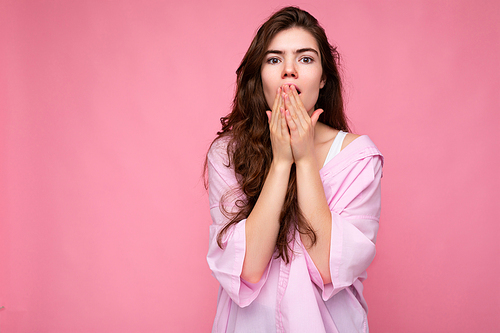 Photo of young shocked amazed surprised winsome nice curly brunette woman with sincere emotions wearing casual pink shirt isolated over pink background with copy space and covering mouth with hands.