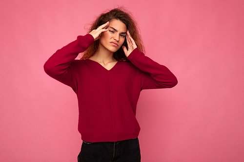 Young emotional attractive brunette curly woman with sincere emotions wearing trendy pink pullover isolated on pink background with copy space and having headache.