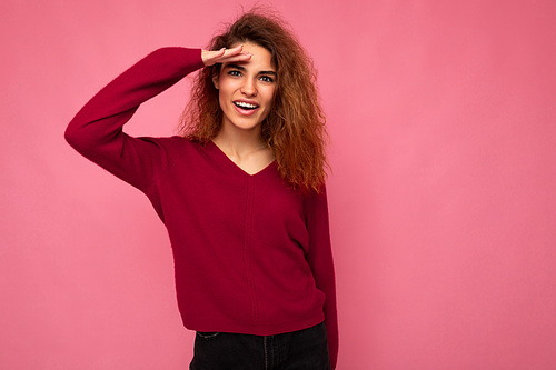 Portrait of young positive happy joyful beautiful brunette curly woman with sincere emotions wearing trendy pink pullover isolated on pink background with copy space and finding something.