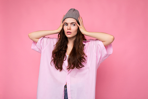 Attractive cute nice adorable tender young curly brunette woman wearing pink shirt and gray hat isolated on pink background with copy space and blocking ears. Hear no evil concept.