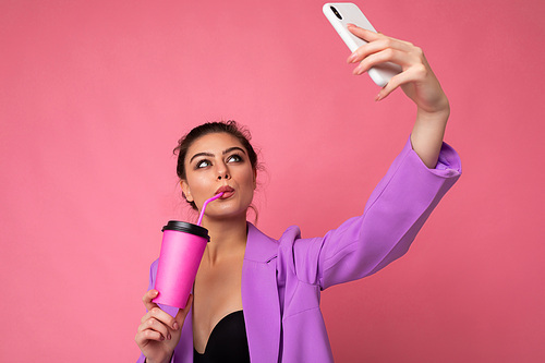 Close-up portrait of a beautiful young brunet woman in fashionable purple suit on a pink background in studio in a holding a popular phone and taking selfie photo and drinking through a straw beverage. Free space