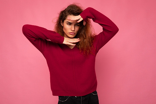 Portrait of young serious beautiful brunette curly woman with sincere emotions wearing trendy pink pullover isolated on pink background with copy space and grimacing with hands.