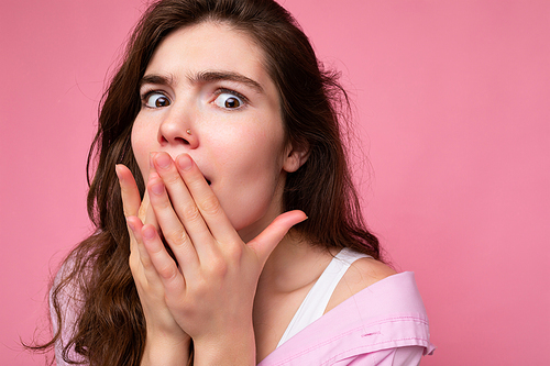 Closeup of young shocked astonished beautiful curly brunet female person with sincere emotions wearing casual pink shirt isolated over pink background with free space and covering mouth with hands.