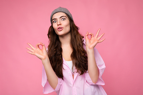 Portrait of young positive surprised beautiful curly brunette woman with sincere emotions wearing trendy pink shirt and grey hat isolated on pink background with empty space and showing ok gesture. It's good.