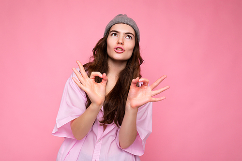 Portrait of young positive emotional funny attractive curly brunette woman with sincere emotions wearing stylish pink shirt and grey hat isolated on pink background with copy space and showing okay gesture. It's fine.