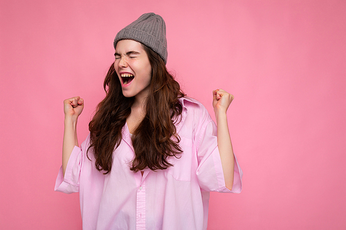 Beautiful happy positive young curly brunette woman wearing pink shirt and grey hat isolated on pink background with copy space shouting yeah..