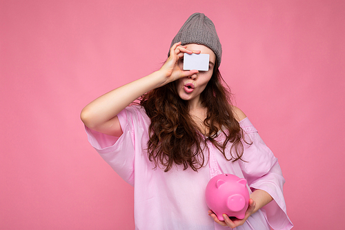 Beautiful amazed surprised young brunette woman wearing shirt isolated on pink background with free space and holding pink pig money box and credit card for mockup.