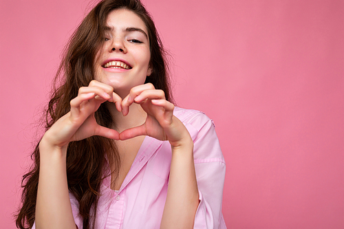 Photo of young positive happy smiling beautiful wavy-haired brunette woman with sincere emotions wearing trendy pink shirt isolated over pink background with free space and showing heart shape with hands.