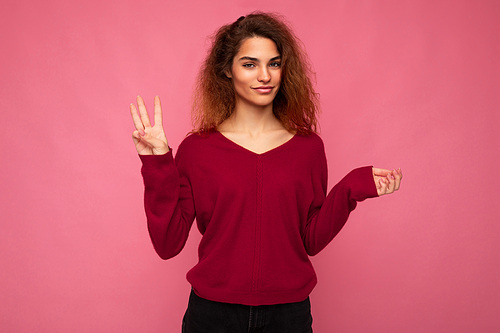 Photo of young positive attractive brunette wavy lady with sincere emotions wearing casual pink sweater isolated over pink background with copy space and showing three fingers.