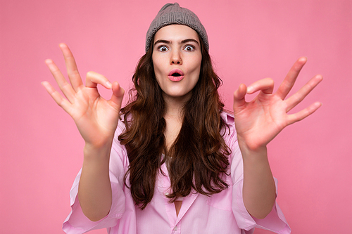 Portrait of young positive shocked amazed attractive curly brunette woman with sincere emotions wearing stylish pink shirt and grey hat isolated on pink background with copy space and showing okay gesture. Wow concept.
