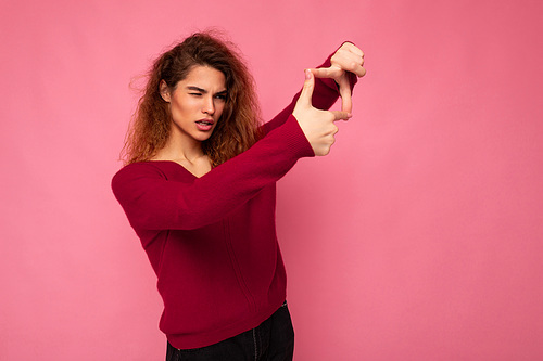 Portrait of young positive pretty brunette curly woman with sincere emotions wearing trendy pink sweater isolated on pink background with copy space and showing camera gesture with hands taking photo.
