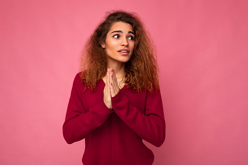 Photo of young emotional beautiful brunette curly woman with sincere emotions wearing trendy pink pullover isolated over pink background with free space and praying with hands holding together askinf for forgiveness.