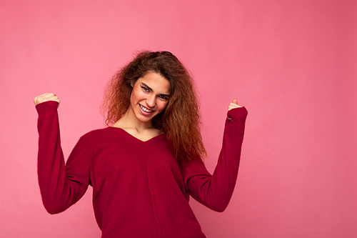 Photo of young emotional happy positive charming brunette curly lady with sincere emotions wearing casual pink pullover isolated over pink background with copy space and celebrating winning.