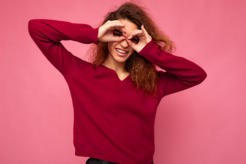 Portrait of young positive happy joyful beautiful brunette curly woman with sincere emotions wearing trendy pink pullover isolated on pink background with copy space and showing glasses gesture with hands.